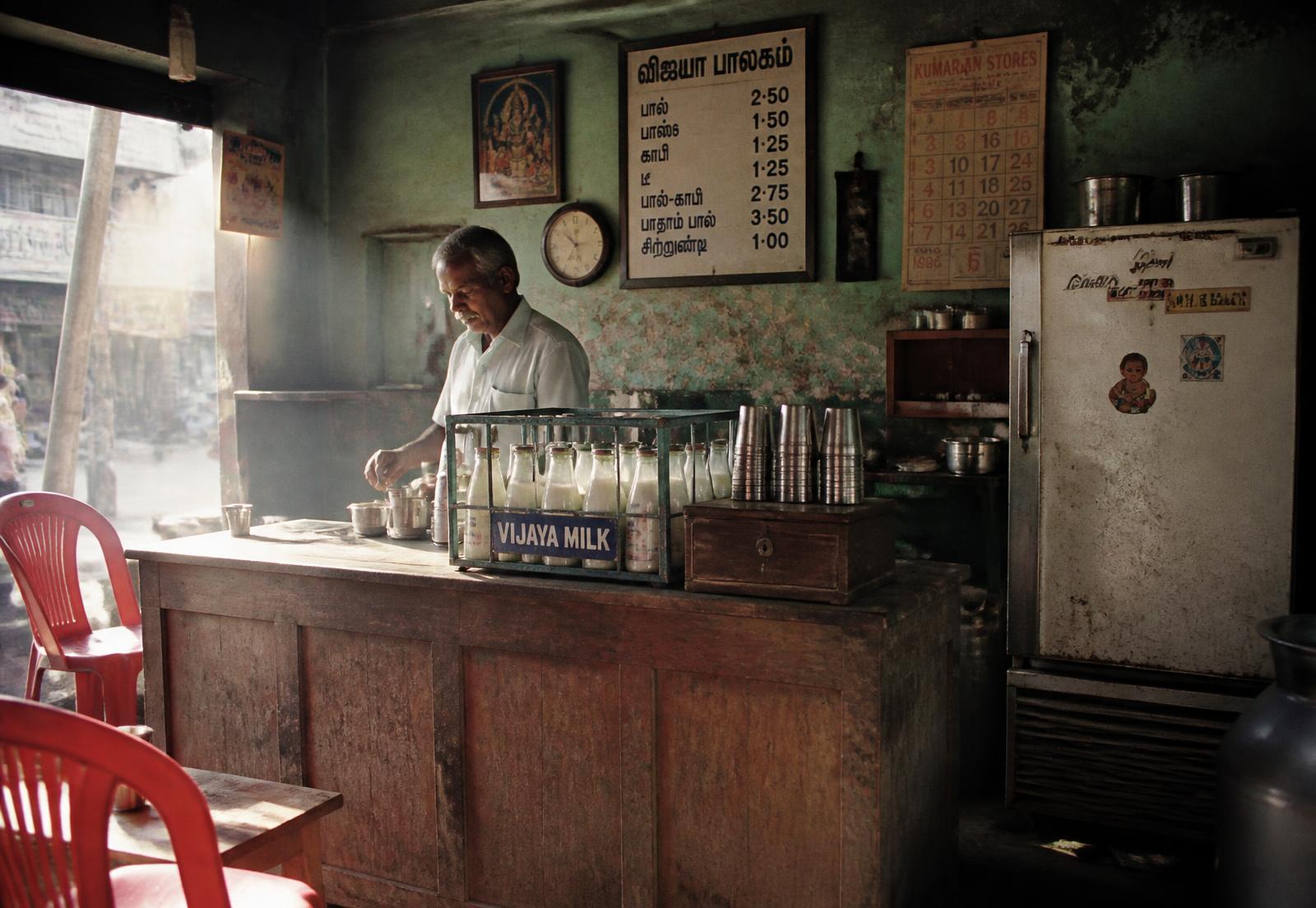 Inside a 1990s South Indian milk shop: wooden counter, crate of glass bottles, steel tumblers, old refrigerator and a handwritten price board